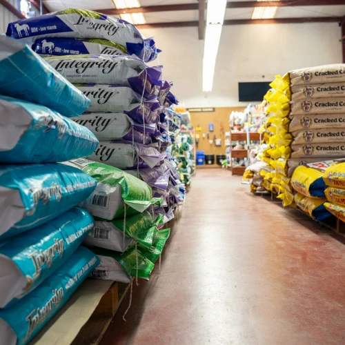 A brightly lit warehouse aisle lined with colorful stacked feed and grain bags, with an open path leading toward shelves in the distance. The scene feels orderly and practical.