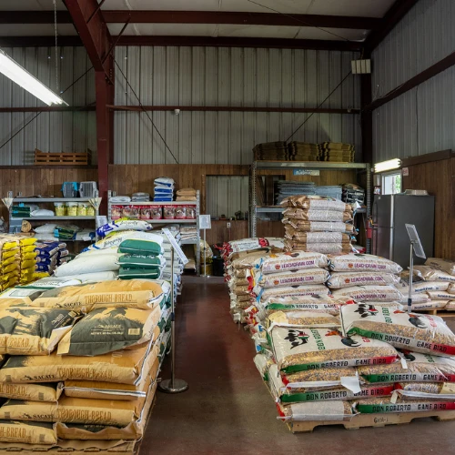 Rows of stacked grain, feed, and fertilizer bags fill a metal-sided warehouse, with shelves and pallets in the background; the orderly store feels busy and practical.