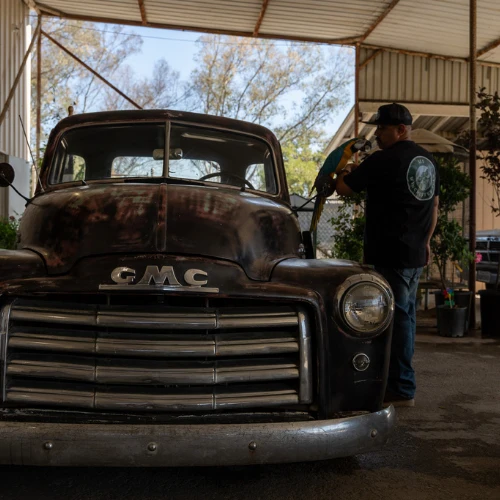 A man in a cap inspects a weathered vintage GMC pickup beneath a metal-roofed workshop, evoking a quiet, hands-on restoration scene.