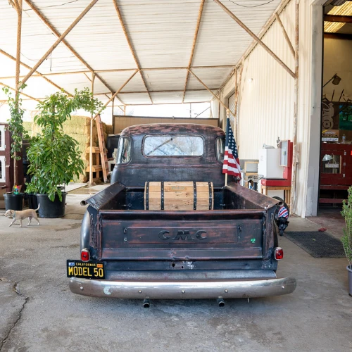 Rear view of a weathered vintage GMC pickup with wooden bed slats, parked in a bright workshop beside an American flag; rustic, nostalgic mood.