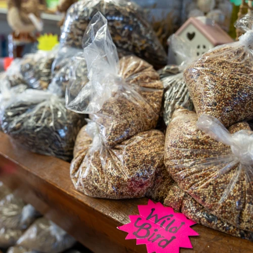 Clear plastic bags of mixed birdseed crowd a wooden market counter, with a bright pink “Wild Bird” label; the abundant display feels rustic and inviting.