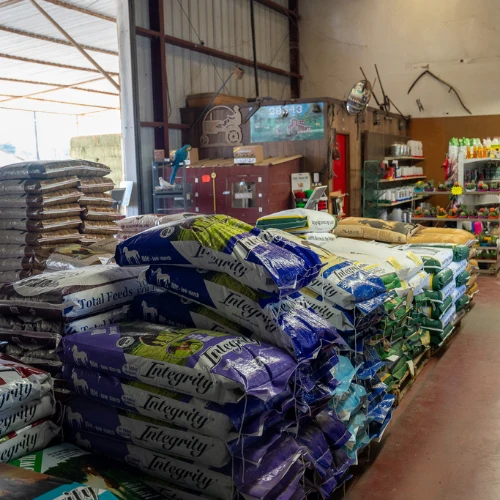 Stacks of purple, white, and green animal-feed bags fill a busy warehouse shop, with shelves of supplies behind them and daylight streaming through an open bay.